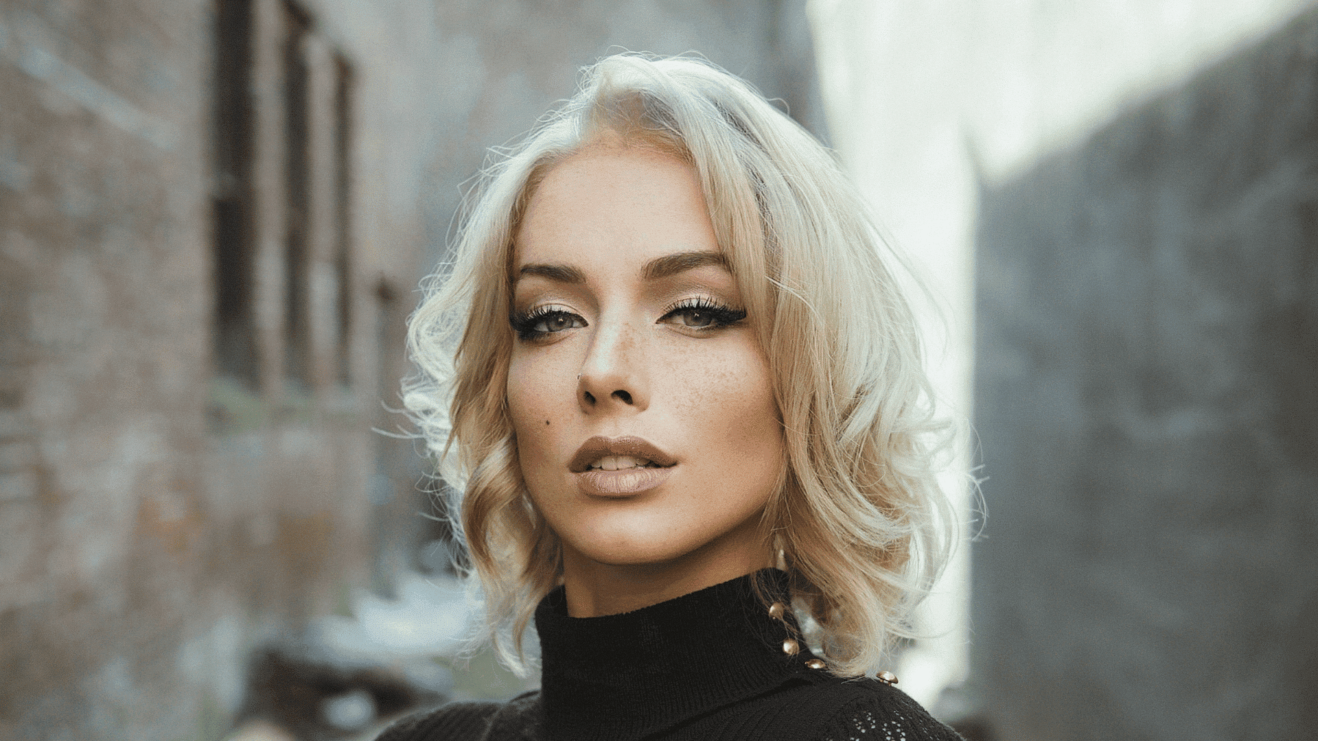 Blonde woman in a black turtleneck standing in an alleyway with a confident expression.