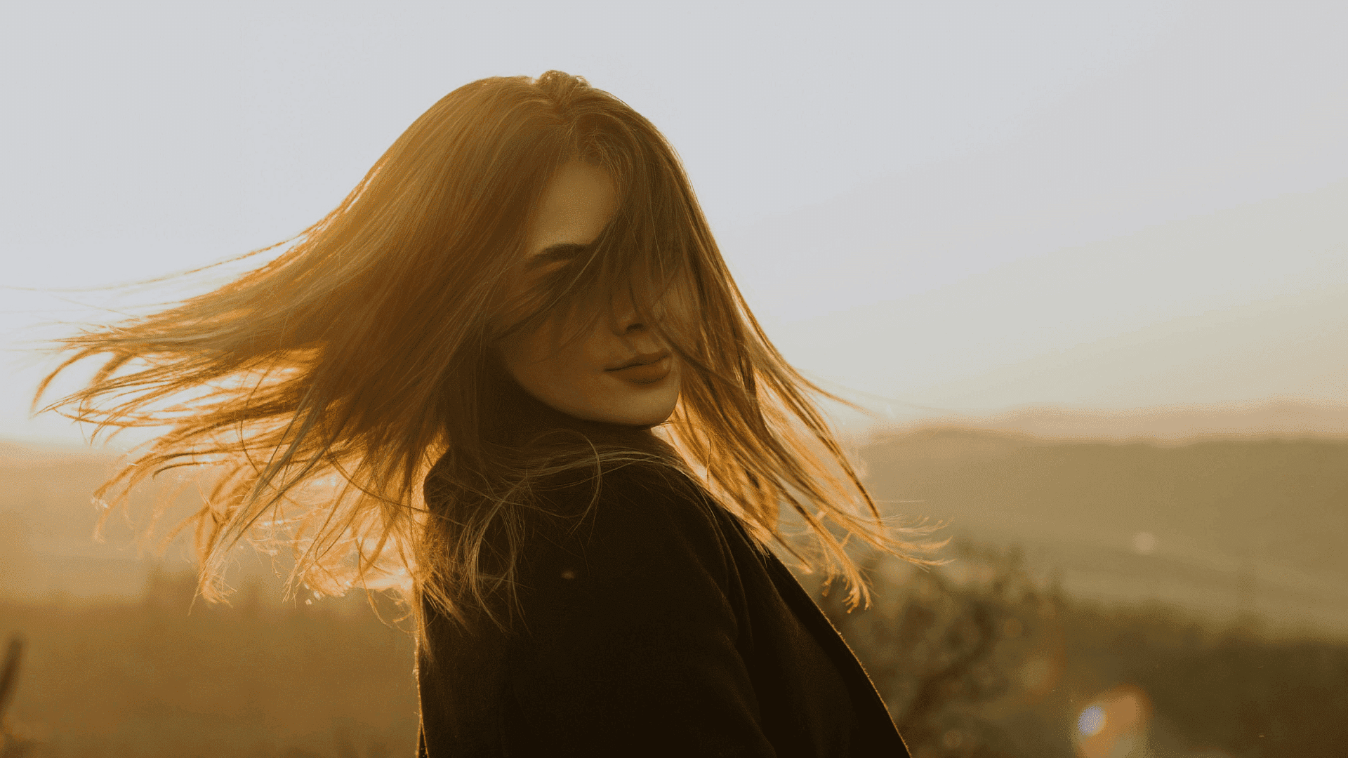 Woman with flowing hair in sunset light, gazing softly with a serene landscape behind.